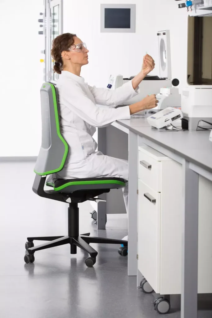 Personne en blouse assise sur un siège NEON à liseré vert, manipulant un instrument sur la paillasse